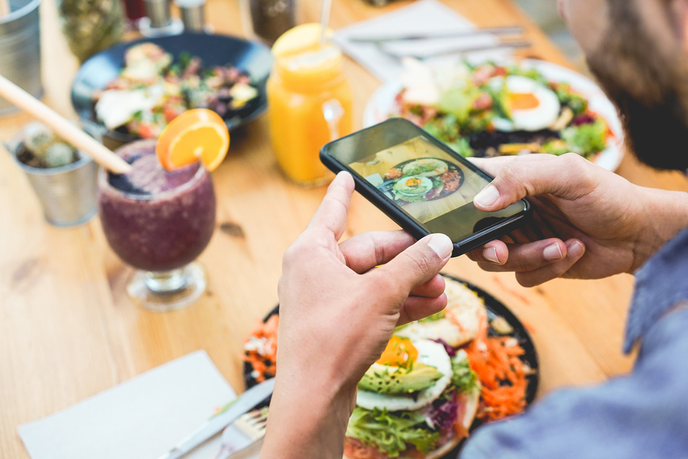 Influencer man eating brunch while making video and photos of dish with mobile phone